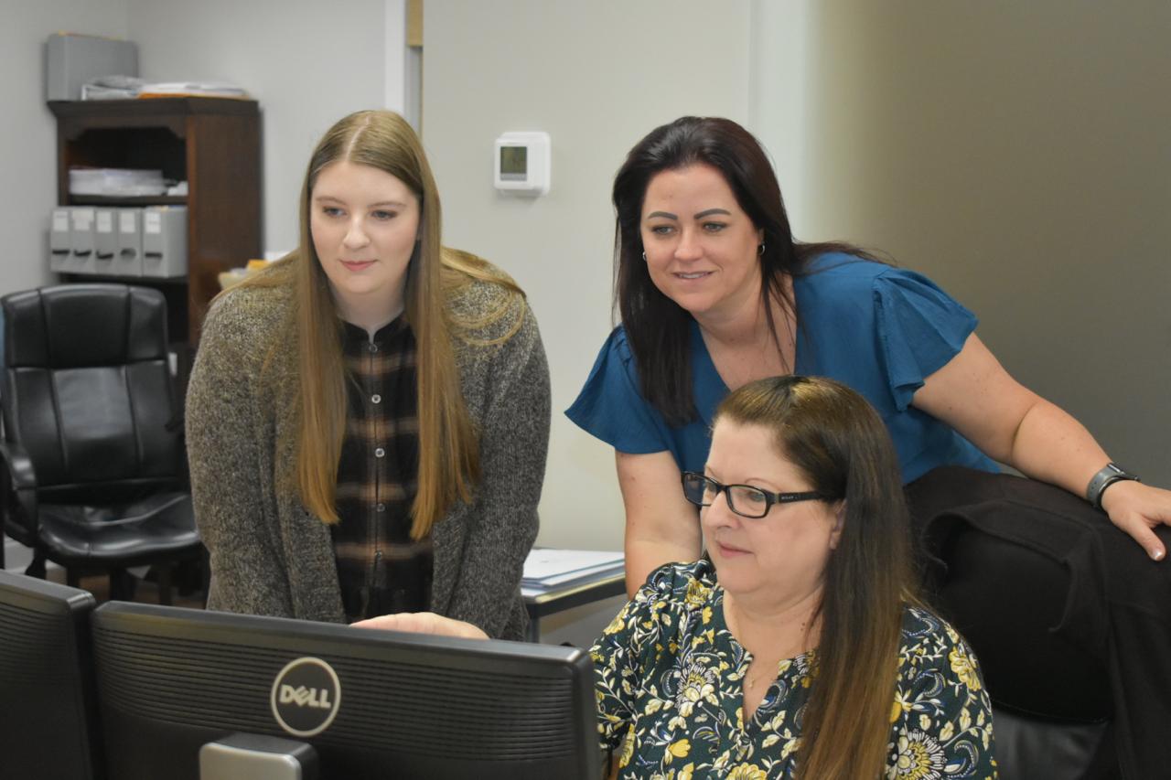 Three women in an office collaboratively examining a computer screen, engaged in discussion and teamwork.